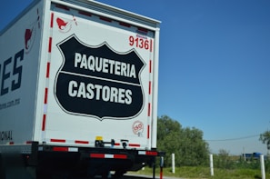 The rear view of a delivery truck with the label 'PAQUETERIA CASTORES' displayed prominently on a black emblem on the back door. The truck has the number '9136' and an 'ISO' certification badge. The background shows a clear blue sky and some greenery.