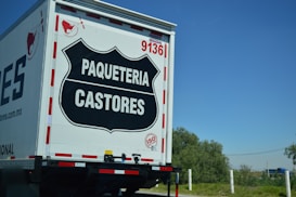The rear view of a delivery truck with the label 'PAQUETERIA CASTORES' displayed prominently on a black emblem on the back door. The truck has the number '9136' and an 'ISO' certification badge. The background shows a clear blue sky and some greenery.