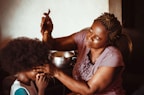 A woman with braided hair is carefully styling a child's hair using a comb. The child is sitting patiently, and there's a focus on the intimate, nurturing interaction between them. The lighting is warm and soft, creating a cozy and homely atmosphere.