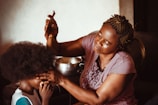 A woman with braided hair is carefully styling a child's hair using a comb. The child is sitting patiently, and there's a focus on the intimate, nurturing interaction between them. The lighting is warm and soft, creating a cozy and homely atmosphere.
