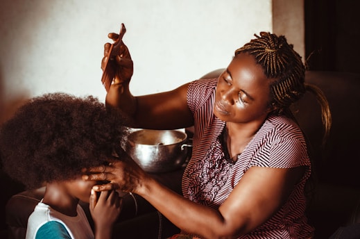 A woman with braided hair is carefully styling a child's hair using a comb. The child is sitting patiently, and there's a focus on the intimate, nurturing interaction between them. The lighting is warm and soft, creating a cozy and homely atmosphere.