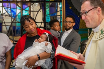 A group of people gathered in front of a colorful stained glass window. A woman is holding a baby dressed in white, cradling it tenderly. A person wearing ecclesiastical attire is holding a book, possibly performing a ceremony. Other individuals are observing, including a man holding a child.