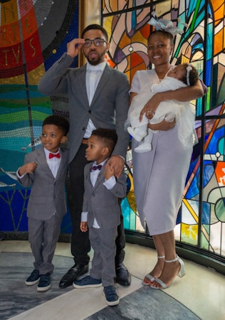 A family poses for a photo in front of a colorful stained glass background. The family consists of two adults and three children. The adults are dressed in formal attire, with the man adjusting his glasses and the woman holding a baby. The two older children are also in suits, looking attentively at the camera.