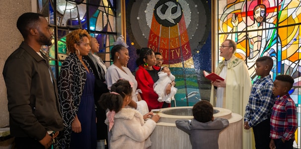 A group of people gathered in a church, participating in a baptism ceremony. An officiant wearing liturgical vestments is speaking, holding a red book. A woman in a red outfit holds a baby dressed in white. Stained glass windows with religious art and symbols are in the background. Children and adults stand around the baptismal font.
