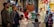 A group of people gathered in a church, participating in a baptism ceremony. An officiant wearing liturgical vestments is speaking, holding a red book. A woman in a red outfit holds a baby dressed in white. Stained glass windows with religious art and symbols are in the background. Children and adults stand around the baptismal font.