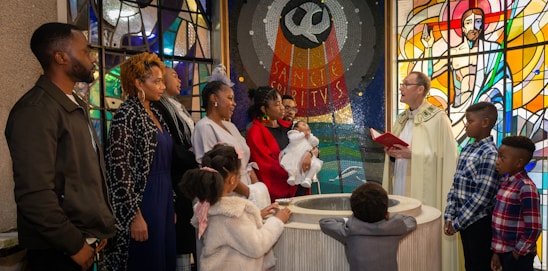 A group of people gathered in a church, participating in a baptism ceremony. An officiant wearing liturgical vestments is speaking, holding a red book. A woman in a red outfit holds a baby dressed in white. Stained glass windows with religious art and symbols are in the background. Children and adults stand around the baptismal font.
