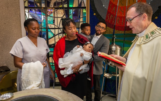 A group of people are gathered around a baptismal font in a church. The focus is on a baby dressed in white, being held by a woman in a red shawl. A priest in liturgical vestments is reading from a book, presumably conducting the baptism ceremony. The background features colorful stained glass windows, adding to the sanctity and beauty of the scene.