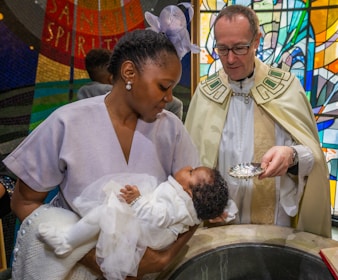 A religious ceremony featuring a woman holding a baby dressed in white, with a priest nearby holding a silver bowl. The setting includes a colorful stained glass background depicting religious symbols.