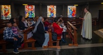 A group of people is seated in a church, listening attentively to a person standing in a robe holding a book. The setting includes wooden pews and colorful stained glass windows in the background.