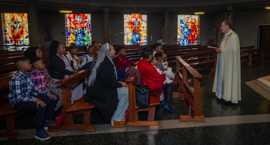 A group of people is seated in a church, listening attentively to a person standing in a robe holding a book. The setting includes wooden pews and colorful stained glass windows in the background.