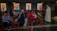 A group of people is seated in a church, listening attentively to a person standing in a robe holding a book. The setting includes wooden pews and colorful stained glass windows in the background.