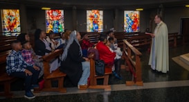 A group of people is seated in a church, listening attentively to a person standing in a robe holding a book. The setting includes wooden pews and colorful stained glass windows in the background.