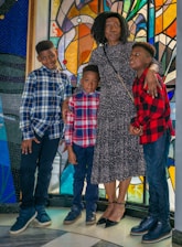 A woman and three children stand closely together in front of a colorful stained glass window. The woman is wearing a dark floral dress and has her arms around the children, who are wearing plaid shirts in shades of blue and red. The setting has vibrant colors and a checkered floor.