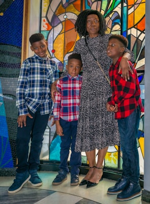 A woman and three children stand closely together in front of a colorful stained glass window. The woman is wearing a dark floral dress and has her arms around the children, who are wearing plaid shirts in shades of blue and red. The setting has vibrant colors and a checkered floor.