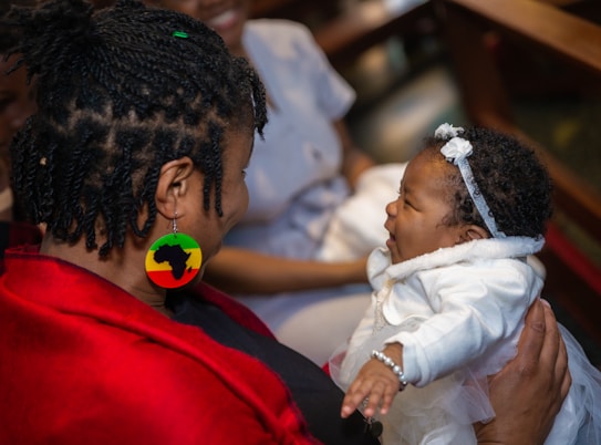 A woman with braided hair wears a colorful circular earring featuring a map silhouette. She is holding a baby dressed in white, who is looking up at her. The baby is wearing a headband and bracelet.