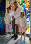 Three children and a baby are standing in front of a vibrant stained glass background. The older child is holding the baby, who is dressed in white, while the two younger children stand on either side, wearing pastel-colored outfits and smiling.