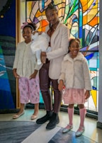 Three children and a baby are standing in front of a vibrant stained glass background. The older child is holding the baby, who is dressed in white, while the two younger children stand on either side, wearing pastel-colored outfits and smiling.