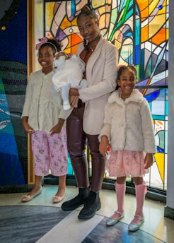 Three children and a baby are standing in front of a vibrant stained glass background. The older child is holding the baby, who is dressed in white, while the two younger children stand on either side, wearing pastel-colored outfits and smiling.