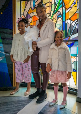 Three children and a baby are standing in front of a vibrant stained glass background. The older child is holding the baby, who is dressed in white, while the two younger children stand on either side, wearing pastel-colored outfits and smiling.