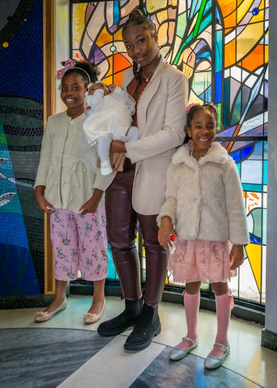 Three children and a baby are standing in front of a vibrant stained glass background. The older child is holding the baby, who is dressed in white, while the two younger children stand on either side, wearing pastel-colored outfits and smiling.