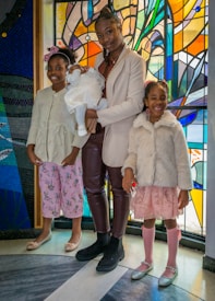 Three children and a baby are standing in front of a vibrant stained glass background. The older child is holding the baby, who is dressed in white, while the two younger children stand on either side, wearing pastel-colored outfits and smiling.