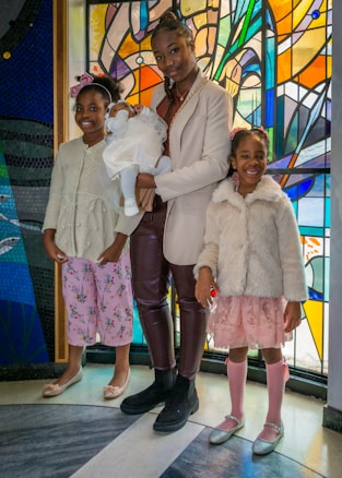 Three children and a baby are standing in front of a vibrant stained glass background. The older child is holding the baby, who is dressed in white, while the two younger children stand on either side, wearing pastel-colored outfits and smiling.