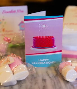 A greeting card featuring a vibrant red cake with a sparkler on top is centered on a marble surface. The card has the words 'Happy Celebrations!' printed on it. Surrounding the card are wrapped candies and possibly other celebratory cards in the background.