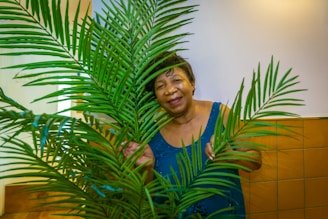 An older woman stands behind a lush green palm plant, smiling warmly and wearing a blue sleeveless top. The background consists of white walls and a section of brown tiles.