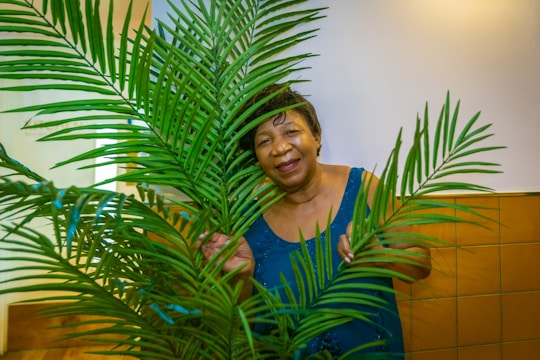 An older woman stands behind a lush green palm plant, smiling warmly and wearing a blue sleeveless top. The background consists of white walls and a section of brown tiles.