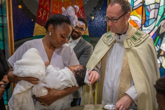 A woman holding a baby is standing next to a man in religious attire. The man is conducting a baptism ceremony, pouring water from a small vessel onto the baby's head. The scene is in a church with colorful stained glass windows in the background.
