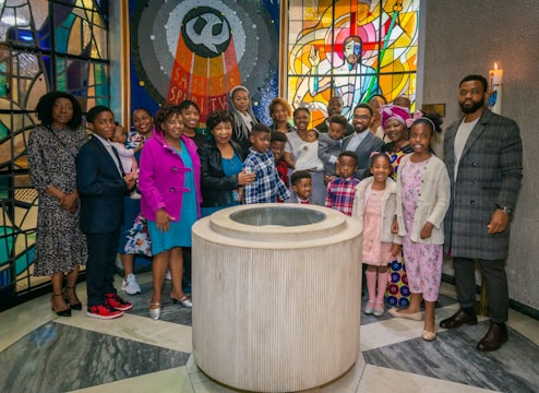 A group of people are gathered around a baptismal font inside a church. They appear to be celebrating an event, possibly a baptism. The setting includes colorful stained glass windows, one depicting a religious figure, and a lit candle on the wall.