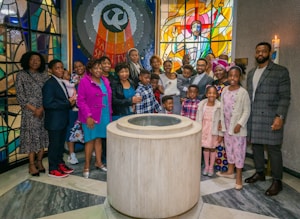 A group of people are gathered around a baptismal font inside a church. They appear to be celebrating an event, possibly a baptism. The setting includes colorful stained glass windows, one depicting a religious figure, and a lit candle on the wall.