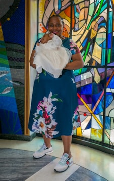 A woman is standing in front of a vibrant stained glass window, holding a baby dressed in a white christening gown. The woman is wearing a floral dress and sneakers. The background features colorful abstract designs and patterns.
