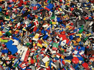 A large collection of assorted colorful Lego bricks and pieces is scattered across the surface. The pieces vary in size and shape, including rectangular blocks, wheels, and specialty bricks, showcasing a vibrant and chaotic mosaic of plastic elements.