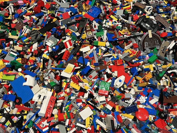 A large collection of assorted colorful Lego bricks and pieces is scattered across the surface. The pieces vary in size and shape, including rectangular blocks, wheels, and specialty bricks, showcasing a vibrant and chaotic mosaic of plastic elements.
