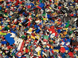 A large collection of assorted colorful Lego bricks and pieces is scattered across the surface. The pieces vary in size and shape, including rectangular blocks, wheels, and specialty bricks, showcasing a vibrant and chaotic mosaic of plastic elements.