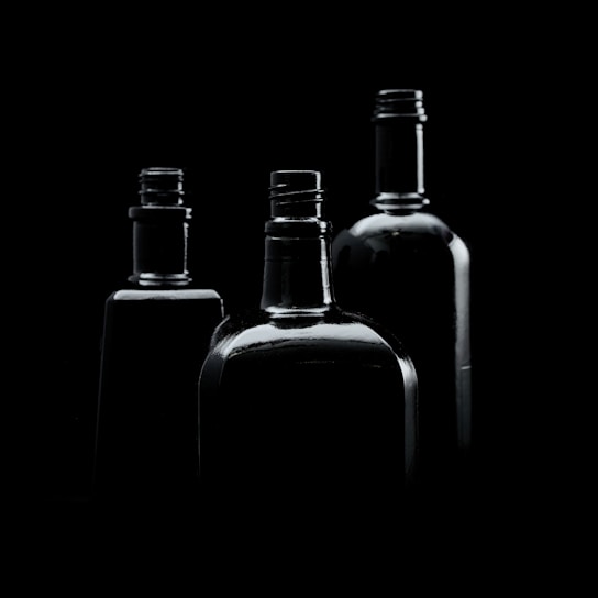 Three dark glass bottles are arranged against a black background. The lighting creates subtle reflections and highlights on the bottles, emphasizing their contours and shapes.