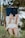A person sitting outdoors in a grassy area holding a blue book titled 'Spiritual Journal' in front of their face. The person is wearing a cream-colored dress and appears to be absorbed in reading. The background consists of blurred trees, suggesting a serene natural setting.