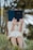 A person sitting outdoors in a grassy area holding a blue book titled 'Spiritual Journal' in front of their face. The person is wearing a cream-colored dress and appears to be absorbed in reading. The background consists of blurred trees, suggesting a serene natural setting.