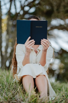 A person sitting outdoors in a grassy area holding a blue book titled 'Spiritual Journal' in front of their face. The person is wearing a cream-colored dress and appears to be absorbed in reading. The background consists of blurred trees, suggesting a serene natural setting.