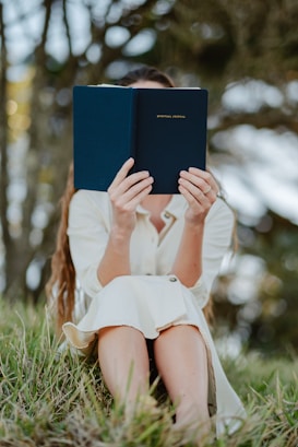 A person sitting outdoors in a grassy area holding a blue book titled 'Spiritual Journal' in front of their face. The person is wearing a cream-colored dress and appears to be absorbed in reading. The background consists of blurred trees, suggesting a serene natural setting.