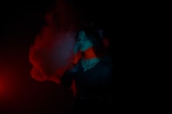 A person is standing with a contemplative expression surrounded by colored smoke. The lighting is dramatic, with red and blue hues casting shadows and highlights.