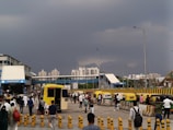 A bustling urban scene with people walking in various directions. In the background, modern high-rise buildings are visible under a dramatic cloudy sky. There is an ambulance parked near the center, and several autorickshaws are lined up on the side. The area appears to be a transit point or station, with covered walkways and traffic barriers.