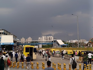 A bustling urban scene with people walking in various directions. In the background, modern high-rise buildings are visible under a dramatic cloudy sky. There is an ambulance parked near the center, and several autorickshaws are lined up on the side. The area appears to be a transit point or station, with covered walkways and traffic barriers.