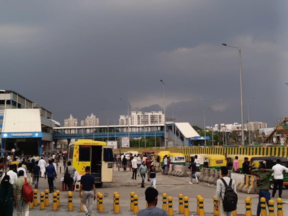 A bustling urban scene with people walking in various directions. In the background, modern high-rise buildings are visible under a dramatic cloudy sky. There is an ambulance parked near the center, and several autorickshaws are lined up on the side. The area appears to be a transit point or station, with covered walkways and traffic barriers.