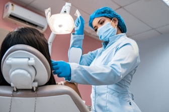 A dental professional in a blue uniform and mask is attending to a patient in a dental chair. The dentist is using dental equipment under a bright examination light, while the room has a calm ambiance with pastel-colored walls.