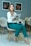 A woman wearing a white blazer and teal pants is seated on a modern chair, holding a magazine titled 'Vida e Saúde'. There are framed floral artworks on the wall in the background and additional chairs arranged nearby. She has a calm and confident expression while a small cup is placed on the magazine she's holding.