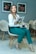 A woman wearing a white blazer and teal pants is seated on a modern chair, holding a magazine titled 'Vida e Saúde'. There are framed floral artworks on the wall in the background and additional chairs arranged nearby. She has a calm and confident expression while a small cup is placed on the magazine she's holding.