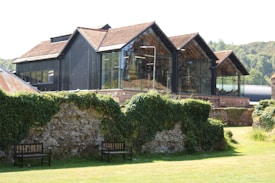 A modern building with large glass windows and black walls, featuring a unique architectural design with multiple pitched roofs. It is surrounded by lush greenery and a stone wall covered in ivy. Two wooden benches are placed against the wall on a well-maintained lawn.