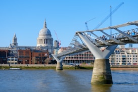 A modern pedestrian bridge extends over a wide river, with a historic cathedral and urban buildings in the background. Several people walk along the bridge, enjoying a clear and sunny day. Construction cranes can be seen above the cityscape, indicating ongoing development or renovation.